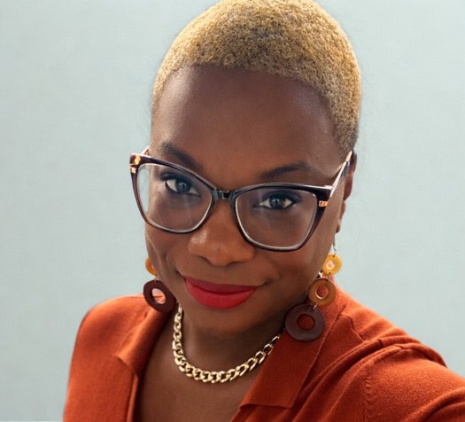 Black woman with golden short hair and black/brown eyeglasses wearing a burn orange top and a gold link chain and earrings with three vertical hollow circles of three shades of brown